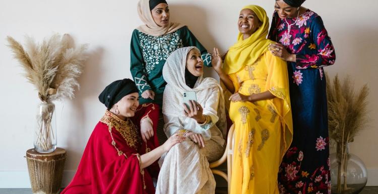 5 women wearing bright-coloured dresses. They are smiling at each other. 