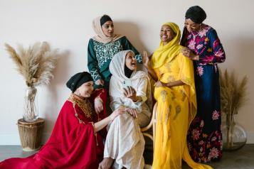 5 women wearing bright-coloured dresses. They are smiling at each other. 